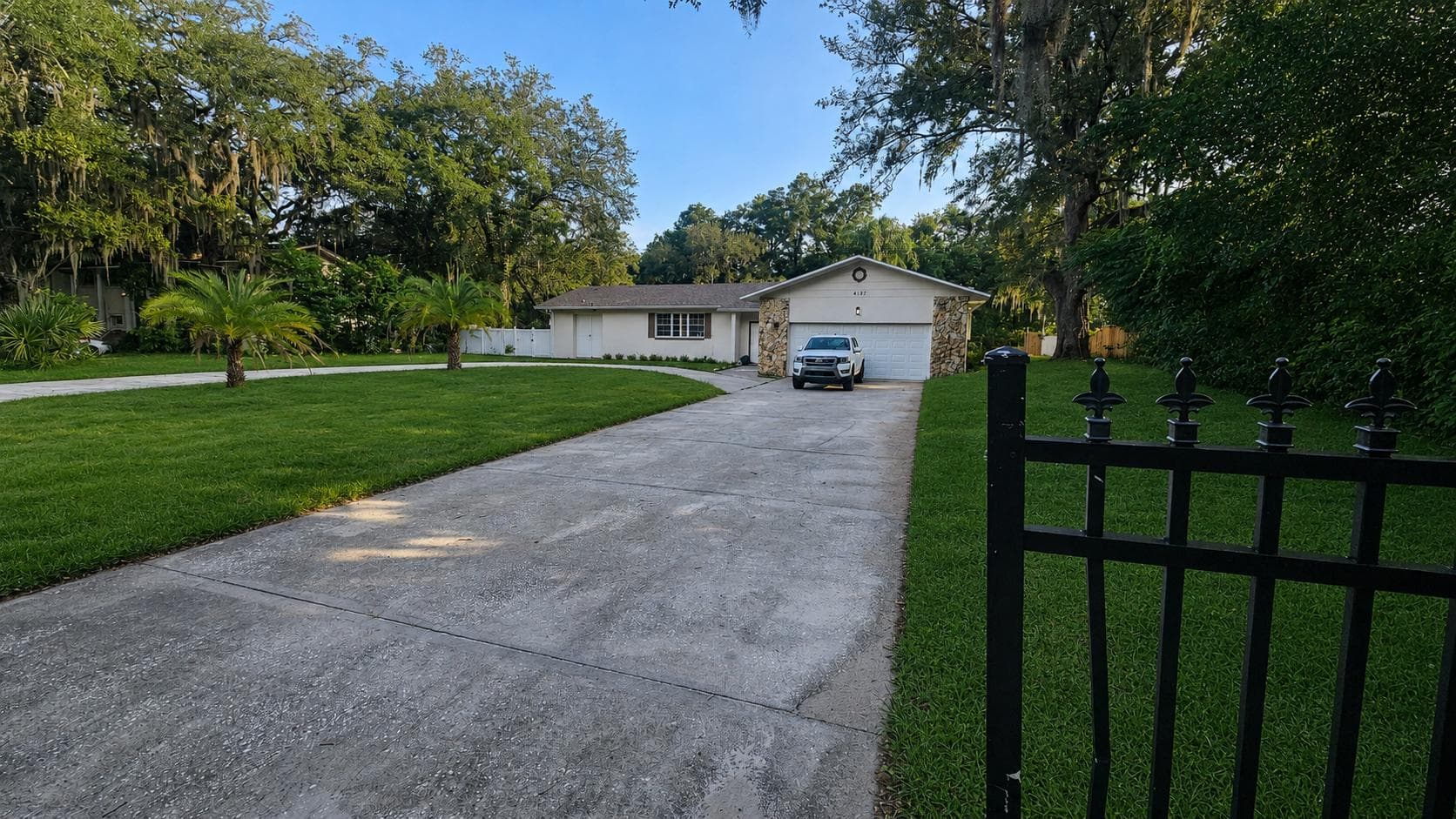 Single-story home with a long driveway, attached garage, and a spacious front lawn shaded by mature trees.